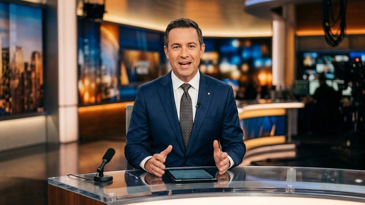 A presenter at a desk in a bright studio.
